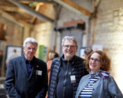 Drei lächelnde Menschen in einem ungezwungenen Innenbereich der Hochschule Coburg. Die beiden Männer tragen Jacken, während die Frau ein gestreiftes Oberteil mit Schal trägt. Im Hintergrund sind eine Backsteinmauer und verschwommene Figuren in der Ferne zu sehen, was zur lebhaften Atmosphäre der Universitätsszene beiträgt.