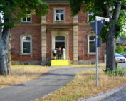 Eine Gruppe von Menschen steht auf den Stufen eines Backsteingebäudes mit Bogenfenstern und einem gelb gestrichenen Eingang, der an den Campus der Hochschule Coburg erinnert. Das Gebäude ist von Bäumen und Gras eingerahmt, in der Nähe gibt es einen Bürgersteig und ein paar geparkte Autos.