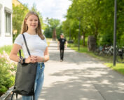 Eine Person mit langen Haaren, bekleidet mit einem weißen Hemd und Jeans, steht auf einem sonnigen Weg in der Nähe der Hochschule Coburg und hält eine schwarze Tasche in der Hand. Im Hintergrund schlendert eine Gestalt in Schwarz spazieren, während Fahrräder entlang des von Grün gesäumten Weges stehen.