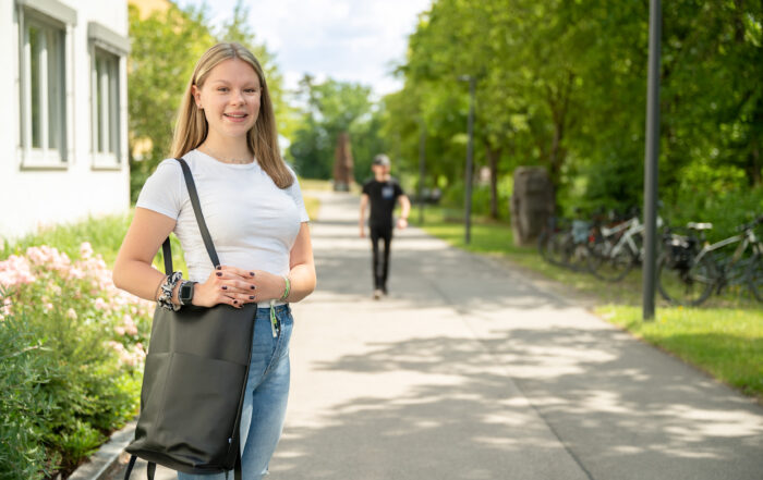 Eine Person mit langen Haaren, bekleidet mit einem weißen Hemd und Jeans, steht auf einem sonnigen Weg in der Nähe der Hochschule Coburg und hält eine schwarze Tasche in der Hand. Im Hintergrund schlendert eine Gestalt in Schwarz spazieren, während Fahrräder entlang des von Grün gesäumten Weges stehen.