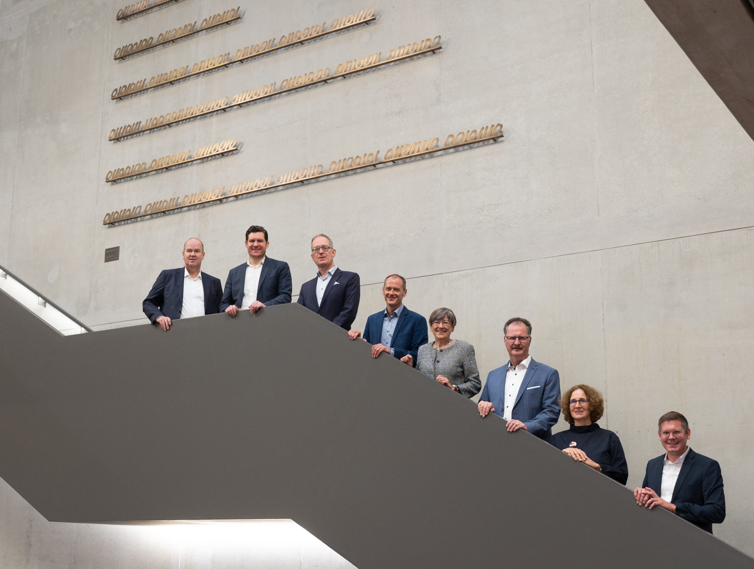 Eine Gruppe von Menschen in formeller Kleidung steht am Rand einer breiten Treppe der Hochschule Coburg, umgeben von einer strukturierten Wand und skulpturaler Textkunst. Sie lächeln und blicken in die Kamera.