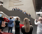 Eine Gruppe von Menschen posiert auf einer Treppe in einem modernen Gebäude der Hochschule Coburg, während mehrere Fotografen und Journalisten den Moment festhalten. Die Kulisse ist in neutralen Tönen gehalten, mit grauen Wänden und Deckenleuchten.