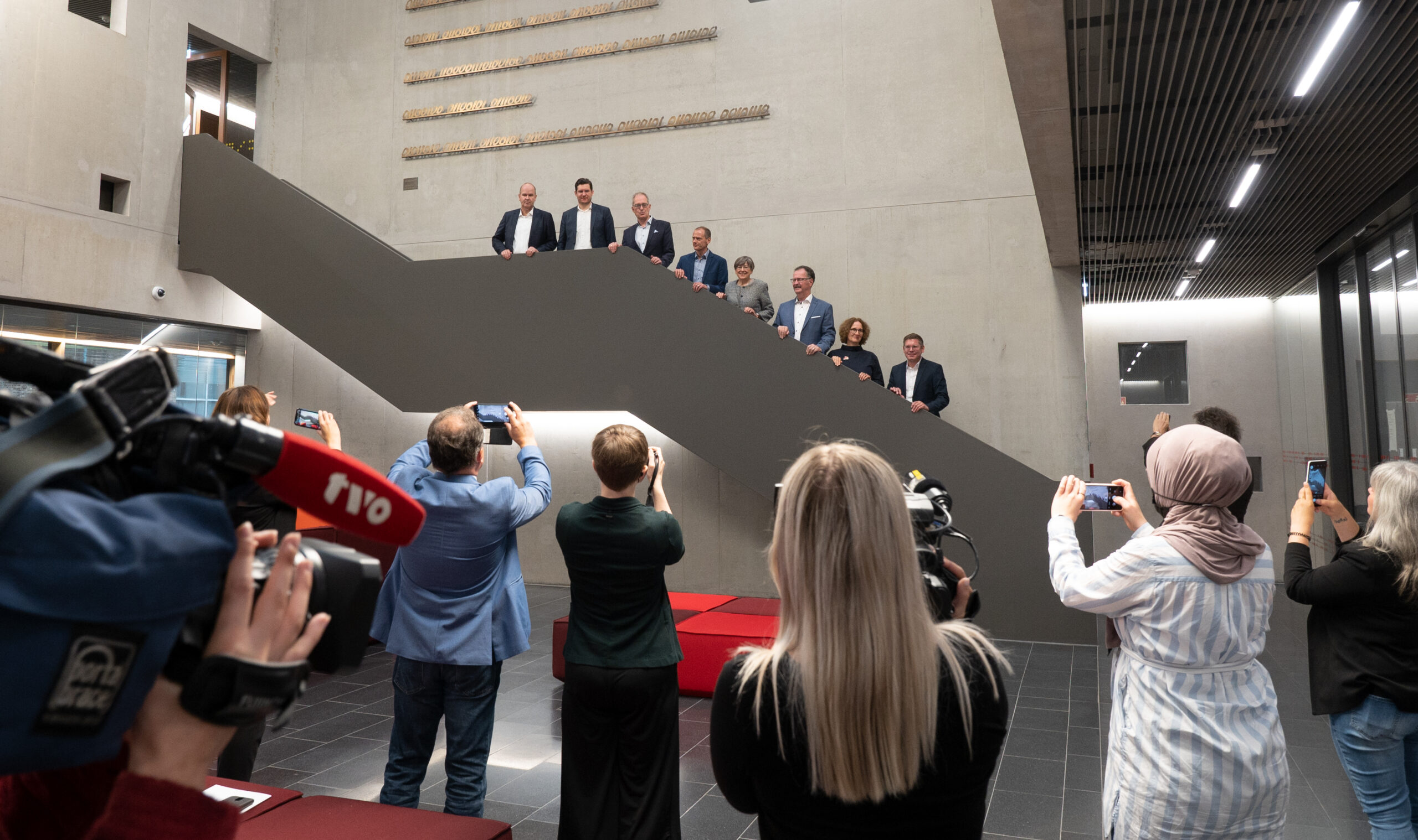 Eine Gruppe von Menschen posiert auf einer Treppe in einem modernen Gebäude der Hochschule Coburg, während mehrere Fotografen und Journalisten den Moment festhalten. Die Kulisse ist in neutralen Tönen gehalten, mit grauen Wänden und Deckenleuchten.