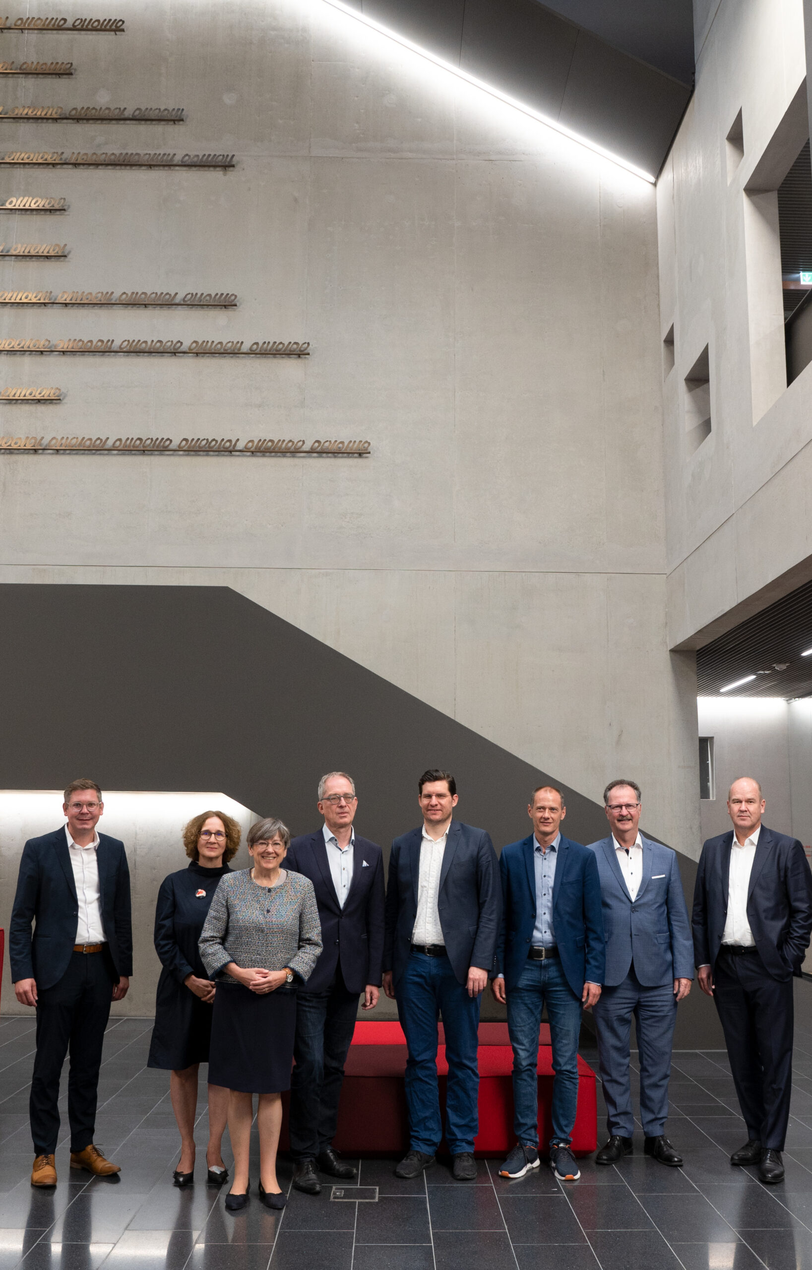 Eine Gruppe von acht Personen in Business-Kleidung steht in einem modernen Gebäude mit hoher Decke der Hochschule Coburg. Sie posieren in zwei Reihen, an der Wand hinter ihnen ist ein Text. Der glänzende Boden reflektiert wunderschön die Deckenbeleuchtung.