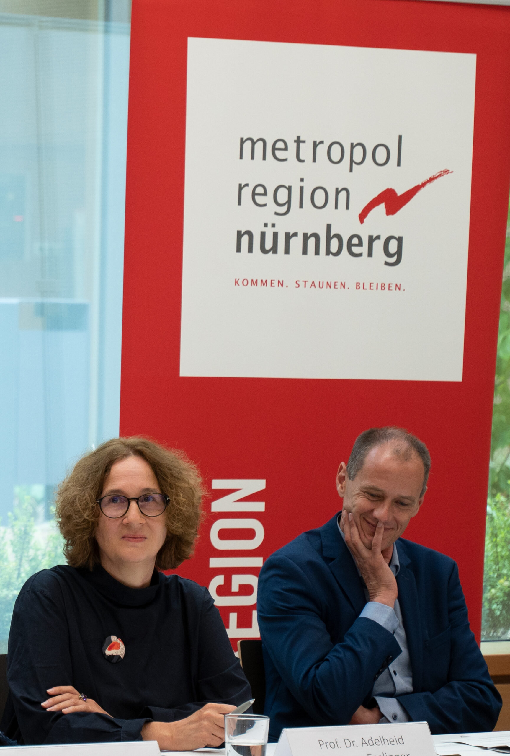 Eine Frau mit lockigem Haar sitzt neben einem Mann im Anzug an einem Tisch. Unter einem rot-weißen Banner steht „Metropolregion Nürnberg“ und der Slogan „kommen. staunen. bleiben“. Auf einem Namensschild steht „Prof. Dr. Adelheid“, die die Hochschule Coburg souverän und repräsentativ vertritt.