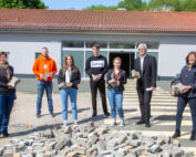 Eine Gruppe von sieben Personen steht vor einem Gebäude der Hochschule Coburg. Jeder hält einen Betonblock. Sie posieren hinter einem verstreuten Haufen von Blöcken auf dem Boden. Das Gebäude hat ein sichtbares Garagentor und ein rotes Ziegeldach.