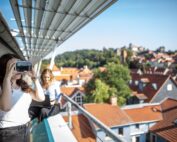 Zwei Personen stehen auf einer Dachterrasse der Hochschule Coburg, im Hintergrund sind rot gekachelte Gebäude und Bäume zu sehen. Eine Person im weißen Hemd trägt ein Virtual-Reality-Headset, während die andere, eine schwarze Jacke tragend, zusieht. Die Szene spielt sich unter einem klaren blauen Himmel ab.