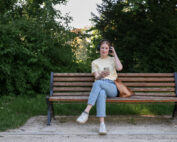 Eine Person mit Kopfhörern und legerer Kleidung, möglicherweise ein Student der Hochschule Coburg, sitzt auf einer Holzbank in einem Park und schaut auf ein Smartphone. Daneben liegt eine braune Tasche. Grünes Laub und Bäume bilden den Hintergrund unter einem klaren Himmel.