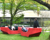 Vier Menschen entspannen auf zwei großen, roten Bänken auf einer von Bäumen umgebenen Rasenfläche der Hochschule Coburg. Im Hintergrund ist ein modernes Gebäude mit großen Fenstern zu sehen. Die Szene ist friedlich und sonnig.