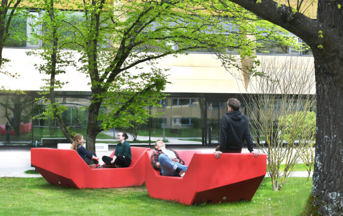 Vier Menschen entspannen auf zwei großen, roten Bänken auf einer von Bäumen umgebenen Rasenfläche der Hochschule Coburg. Im Hintergrund ist ein modernes Gebäude mit großen Fenstern zu sehen. Die Szene ist friedlich und sonnig.
