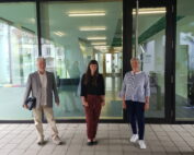 Drei Personen stehen vor dem modernen Gebäude der Hochschule Coburg mit Glastüren. Die Person in der Mitte trägt ein schwarzes Oberteil und rote Hosen; die anderen sind leger gekleidet. Im Hintergrund leuchtet ein Flur mit heller Deckenbeleuchtung, die das moderne Ambiente unterstreicht.