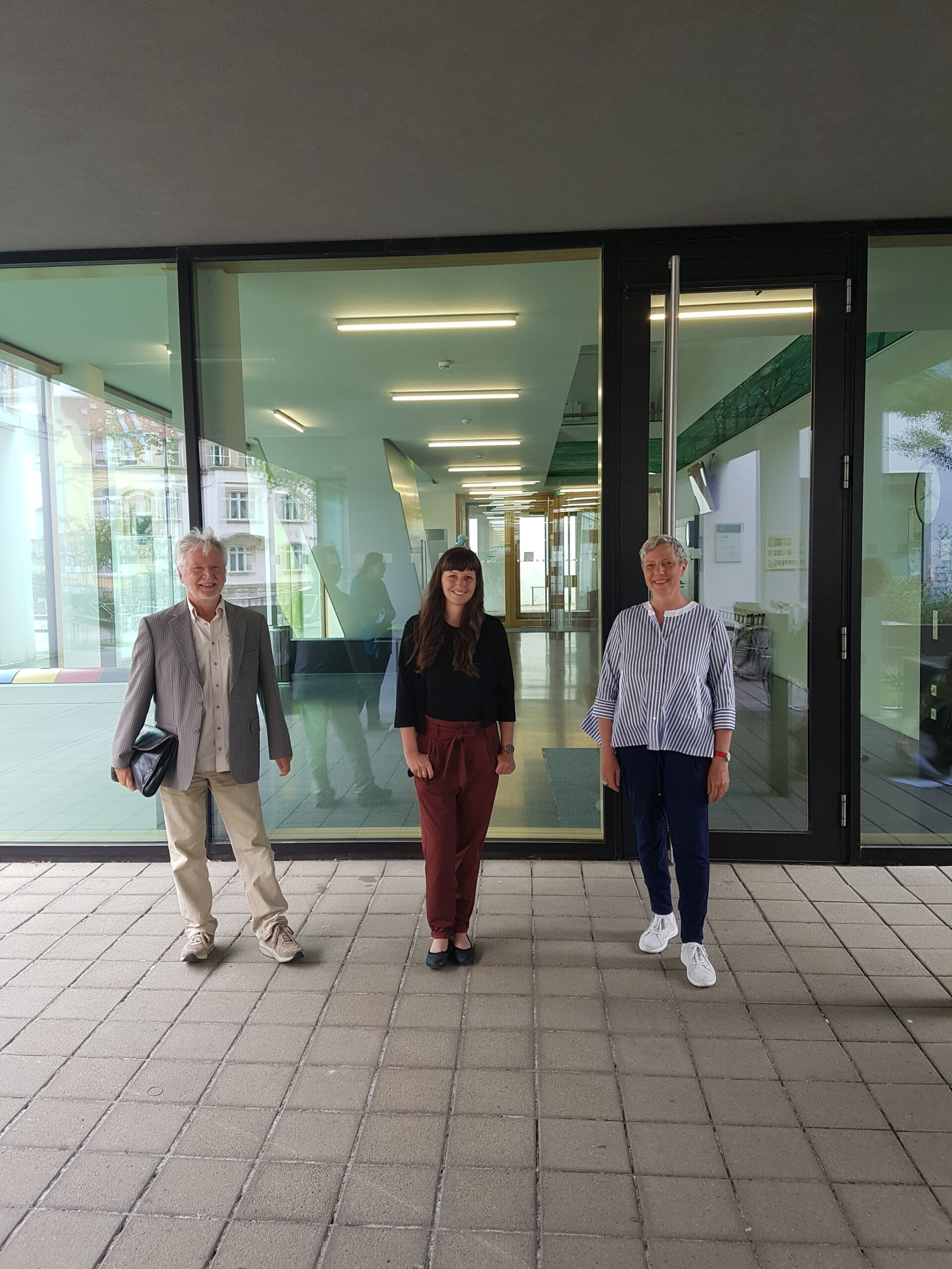 Drei Personen stehen vor dem modernen Gebäude der Hochschule Coburg mit Glastüren. Die Person in der Mitte trägt ein schwarzes Oberteil und rote Hosen; die anderen sind leger gekleidet. Im Hintergrund leuchtet ein Flur mit heller Deckenbeleuchtung, die das moderne Ambiente unterstreicht.