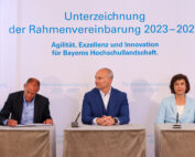 Drei Personen sitzen an einem Tisch unter einem Transparent mit der Aufschrift „Unterzeichnung der Rahmenvereinbarung 2023-2027. Agilität, Exzellenz und Innovation für Bayerns Hochschullandschaft“ während einer Autogrammstunde, die die Zukunft der Hochschule Coburg beleuchtet.