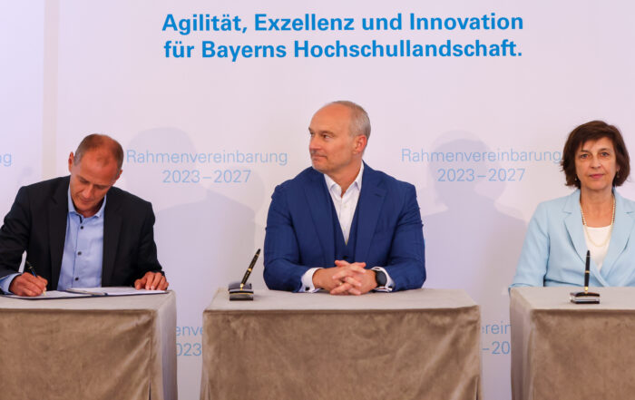 Drei Personen sitzen an einem Tisch unter einem Transparent mit der Aufschrift „Unterzeichnung der Rahmenvereinbarung 2023-2027. Agilität, Exzellenz und Innovation für Bayerns Hochschullandschaft“ während einer Autogrammstunde, die die Zukunft der Hochschule Coburg beleuchtet.