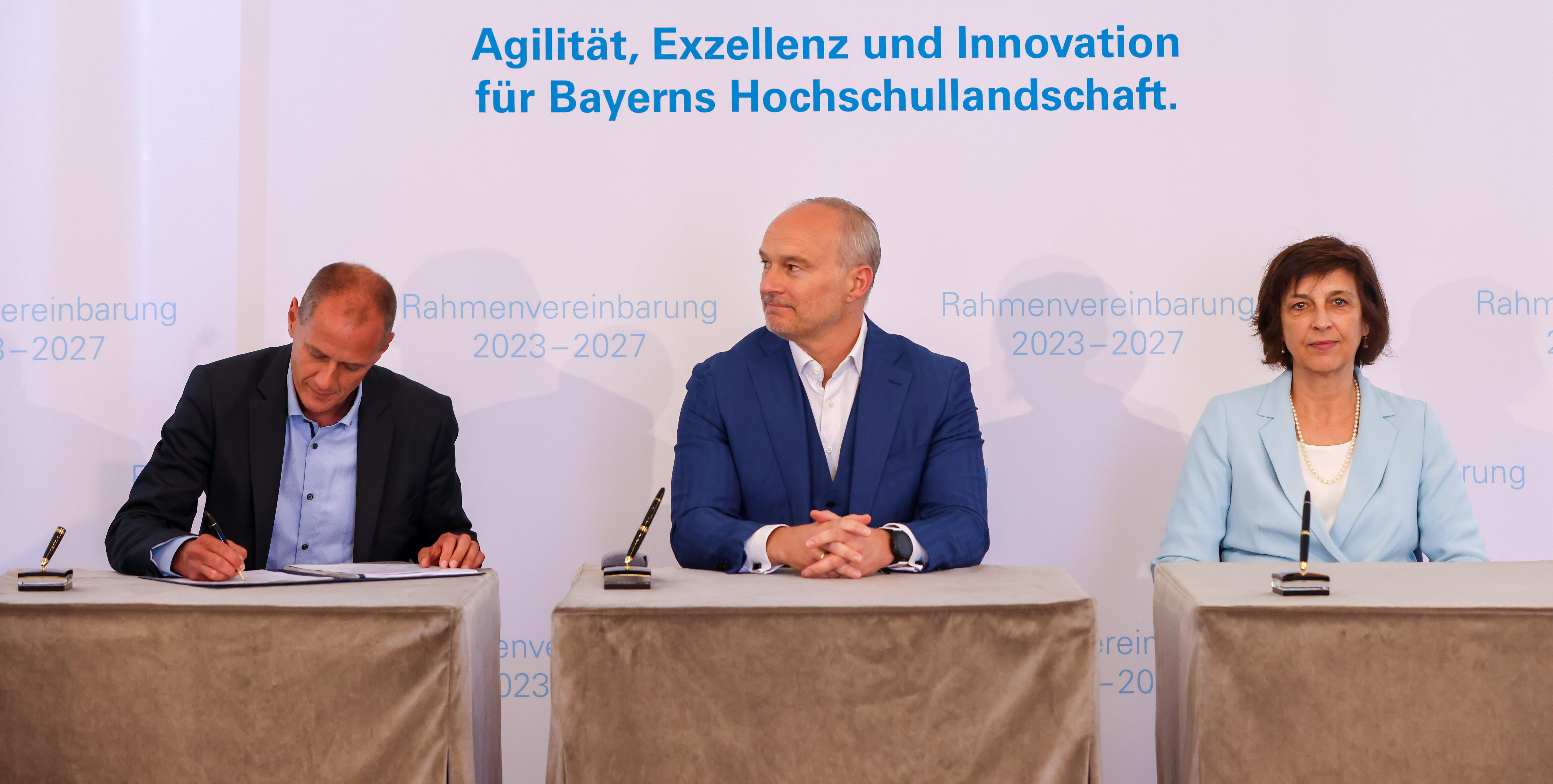 Drei Personen sitzen an einem Tisch unter einem Transparent mit der Aufschrift „Unterzeichnung der Rahmenvereinbarung 2023-2027. Agilität, Exzellenz und Innovation für Bayerns Hochschullandschaft“ während einer Autogrammstunde, die die Zukunft der Hochschule Coburg beleuchtet.