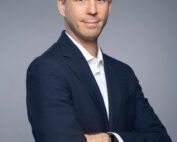 Ein Mann in dunklem Anzug und weißem Hemd, möglicherweise ein Alumnus der Hochschule Coburg, steht mit verschränkten Armen vor einem grauen Hintergrund. Er hat kurze braune Haare, trägt eine Smartwatch am Handgelenk und blickt mit neutralem Gesichtsausdruck direkt in die Kamera.