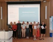 Eine Gruppe von Menschen steht in einem Raum der Hochschule Coburg. Im Hintergrund läuft auf einem Bildschirm eine Abschlusspräsentation. Einige halten stolz Zertifikate in den Händen, alle lächeln und schaffen eine herzliche und professionelle Atmosphäre.