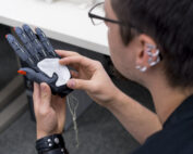 Eine Person der Hochschule Coburg hält eine Handprothese mit roten Fingerspitzen und daran befestigten Drähten und passt das Gerät an oder untersucht es, wobei sie sich auf die Handfläche konzentriert. Sie trägt ein schwarzes Hemd, sichtbare Piercings und ein Lederarmband.
