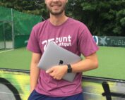 Ein Mann mit Brille, lila T-Shirt und blauen Shorts grinst und hält einen zugeklappten Laptop in der Hand. Er steht vor einer mit Graffiti bedeckten Wand auf dem lebendigen Campus der Hochschule Coburg, im Hintergrund ein grüner Sportplatz und Bäume.