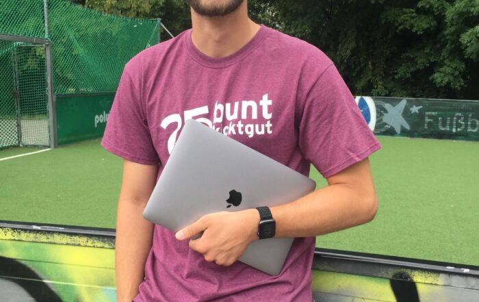Ein Mann mit Brille, lila T-Shirt und blauen Shorts grinst und hält einen zugeklappten Laptop in der Hand. Er steht vor einer mit Graffiti bedeckten Wand auf dem lebendigen Campus der Hochschule Coburg, im Hintergrund ein grüner Sportplatz und Bäume.