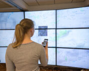 Eine Frau steht vor einer großen Wandtafel mit digitalen Karten an der Hochschule Coburg. Sie hält ein Smartphone in der Hand und betrachtet die Karten auf mehreren Bildschirmen, die Straßen und geografische Merkmale zeigen.