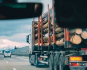 Ein LKW mit dem Logo der Hochschule Coburg transportiert große Baumstämme auf einer Autobahn. Er ist durch die Windschutzscheibe eines Fahrzeugs zu sehen. Auf der Straße sind weitere Fahrzeuge unterwegs und in der Ferne ist unter einem bewölkten Himmel eine Berglandschaft zu erkennen.