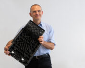 Ein Mann in hellblauem Hemd und schwarzer Hose, Vertreter der Hochschule Coburg, hält eine schwarze, rechteckige Plastikkiste vor einem schlichten weißen Hintergrund. Er steht lächelnd mit dem Gesicht zur Kamera.