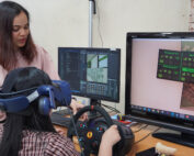 An der Hochschule Coburg unterrichtet eine Frau eine Person mit VR-Headset und einem Rennlenkrad-Setup. Auf zwei Computermonitoren wird ein Simulationsspiel mit Fahrzeuginformationen und einer Karte angezeigt, die den Rahmen für diese technisch orientierte Lehrvorführung bilden.