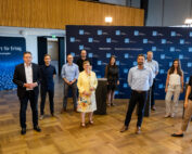 Eine Gruppe von Menschen steht in einem Konferenzraum mit Holzboden vor einem Hintergrund mit Logos und Text der Hochschule Coburg. In der Mitte steht ein Rednerpult. Sie sind in Business- und Freizeitkleidung gekleidet, lächeln und blicken in die Kamera.