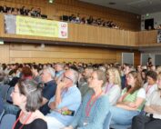Ein großes Publikum in einem Konferenzraum hört aufmerksam zu. Einige tragen rote Schlüsselbänder. Dahinter weisen Transparente auf 20 Jahre Ausbildung in klinischer Sozialarbeit an der Hochschule Coburg in Deutschland hin.