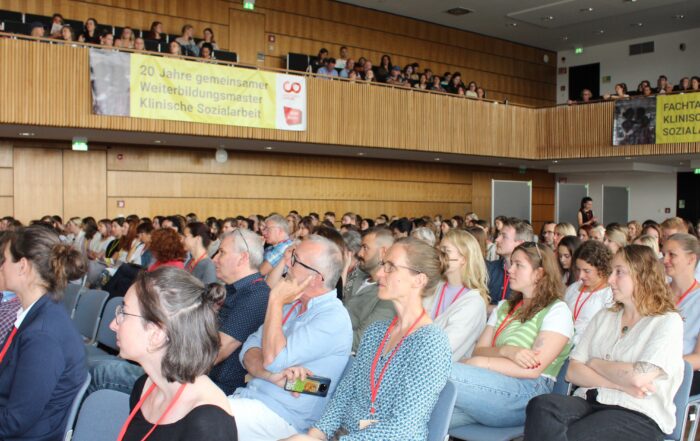 Ein großes Publikum in einem Konferenzraum hört aufmerksam zu. Einige tragen rote Schlüsselbänder. Dahinter weisen Transparente auf 20 Jahre Ausbildung in klinischer Sozialarbeit an der Hochschule Coburg in Deutschland hin.