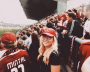 Eine lächelnde Frau mit langen blonden Haaren, einer Brille und einer roten Kappe mit dem Schriftzug „Hochschule Coburg“ steht in der Menge des Stadions. Sie ist von Menschen in Rot und jubelnden Fans umgeben. Die Atmosphäre ist energiegeladen und lebhaft.