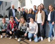 Eine Gruppe von 13 Personen, sechs Männer und sieben Frauen, versammelt sich auf einem Holzdeck vor einem Gebäude der Hochschule Coburg. Sie lächeln herzlich in die Kamera, einige halten Smartphones in den Händen. Der sonnige Tag wirft verspielte Schatten auf den Boden.