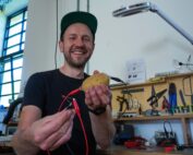 Ein lächelnder Mann mit Mütze präsentiert stolz eine mit Elektronik verkabelte Kartoffel in seiner Werkstatt. In der Hand hält er ein kleines elektronisches Bauteil und steht zwischen Werkzeugen, einer Schreibtischlampe und diversen Gadgets – ein echter Heimwerker von der Hochschule Coburg.