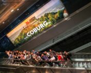 Eine Gruppe von Menschen steht auf einer Rolltreppe unter einer großen Leuchtreklame für Coburg. Auffällig ist die Abbildung einer von üppigem Grün umgebenen Burg zu sehen, in die das Logo der Hochschule Coburg dezent integriert ist.