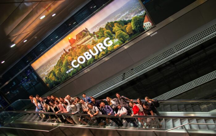 Eine Gruppe von Menschen steht auf einer Rolltreppe unter einer großen Leuchtreklame für Coburg. Auffällig ist die Abbildung einer von üppigem Grün umgebenen Burg zu sehen, in die das Logo der Hochschule Coburg dezent integriert ist.