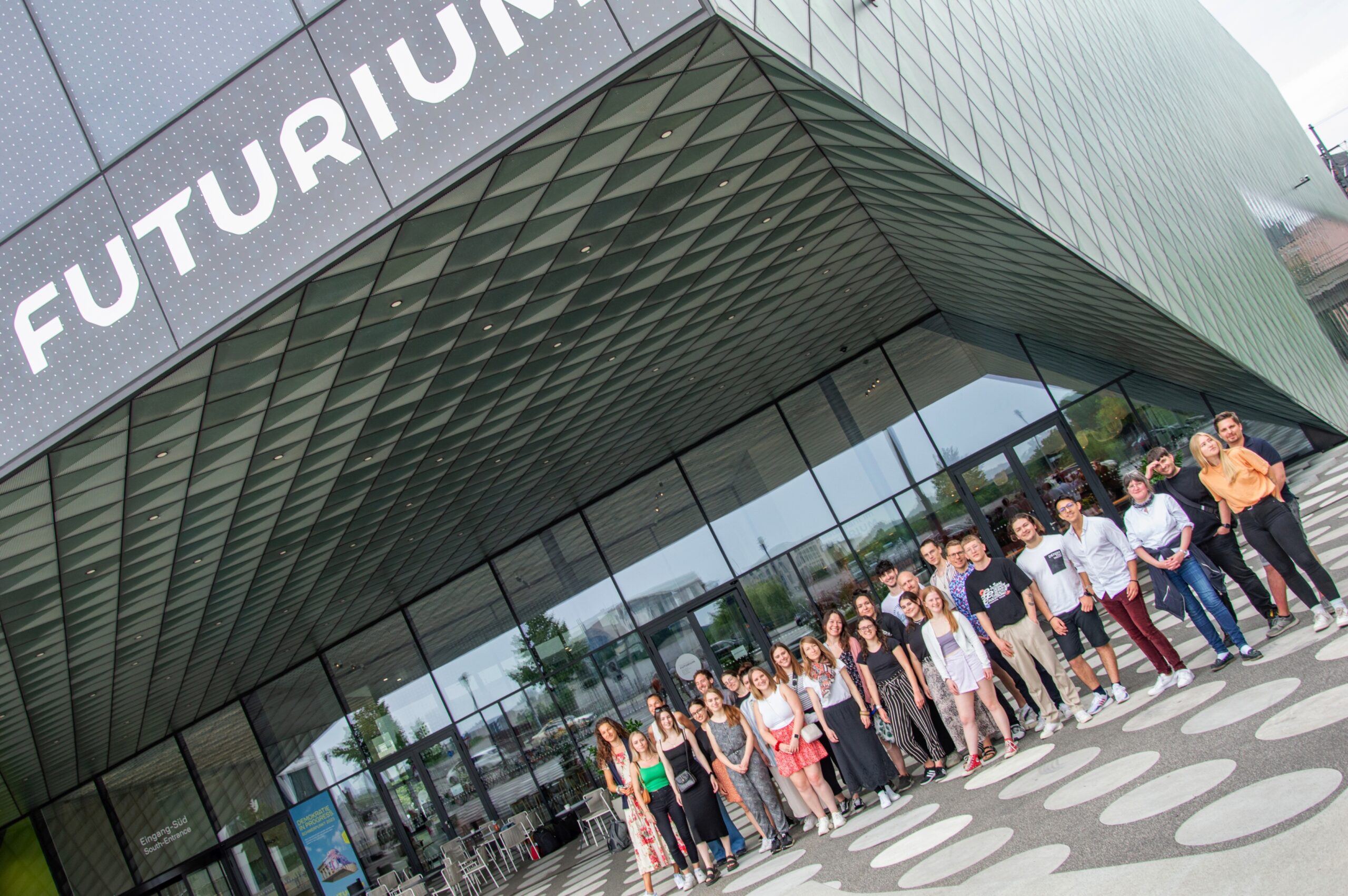 Eine Gruppe von Menschen steht vor dem modernen FUTURIUM-Gebäude, dessen geometrische Glasfassade den gemusterten Dachüberstand wunderschön widerspiegelt. Unter ihnen sind Besucher der Hochschule Coburg, die alle eifrig für ein Erinnerungsfoto posieren.