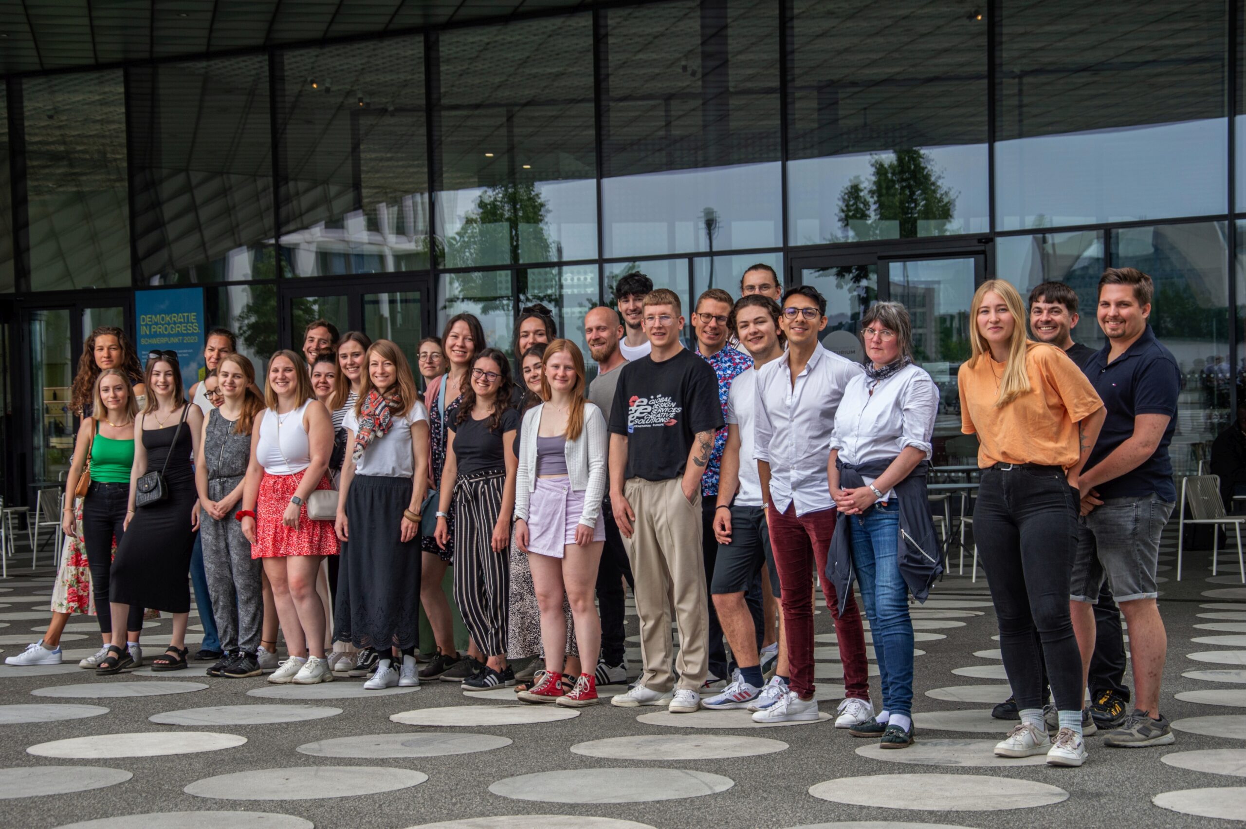 Eine Gruppe von Menschen, einige in Freizeitkleidung, andere eher formell gekleidet, steht gemeinsam vor dem modernen Glasgebäude der Hochschule Coburg. Der Boden ist mit einem kreisförmigen Muster versehen, die Stimmung der Gruppe ist locker und gesellig.