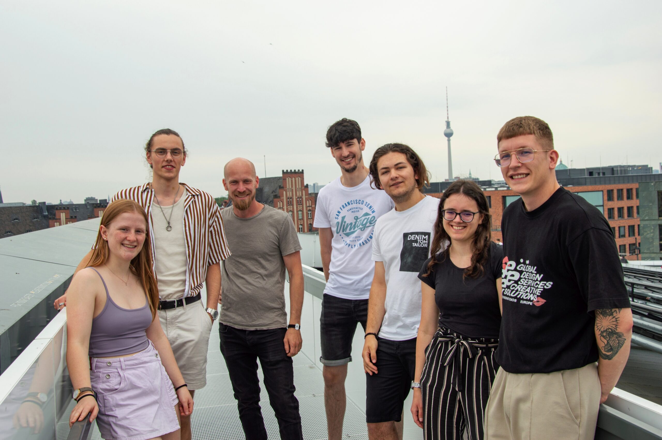Eine Gruppe von sieben jungen Erwachsenen der Hochschule Coburg steht lächelnd auf einem Dach, hinter ihnen erstreckt sich die Stadtlandschaft. Sie wirken entspannt und leger gekleidet vor dem Hintergrund eines bedeckten Himmels.