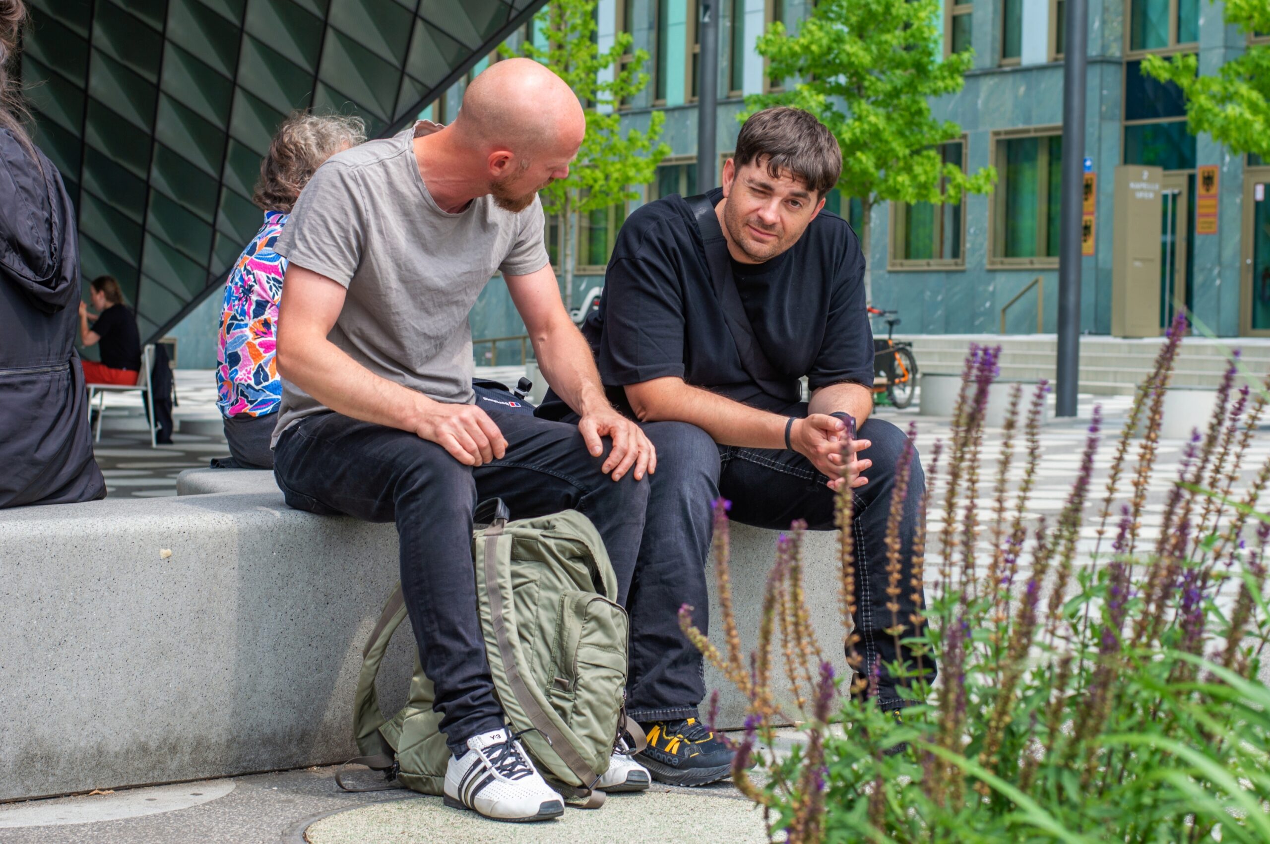 Zwei Männer sitzen auf einer Betonbank im Freien und unterhalten sich in der Nähe der Hochschule Coburg. Einer trägt ein graues Hemd und Jeans, der andere ein schwarzes Hemd und eine schwarze Hose. In der Nähe liegt zwischen modernen Gebäuden und Grün ein grüner Rucksack.