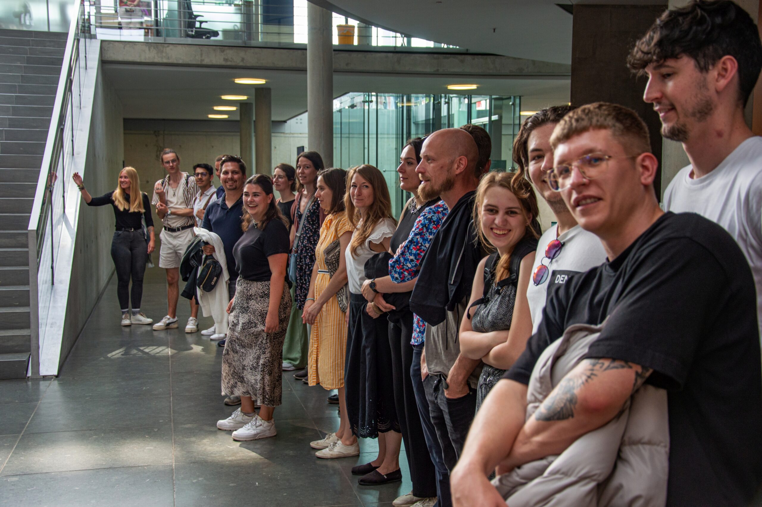 Eine Gruppe von Menschen steht in einem Flur der Hochschule Coburg, gegenüber einer Treppe. Einige lächeln und unterhalten sich mit der Person, die das Foto macht. Die Umgebung wirkt modern, mit Glaswänden und Betonböden. Sie sind leger gekleidet und es sind Männer und Frauen gemischt.