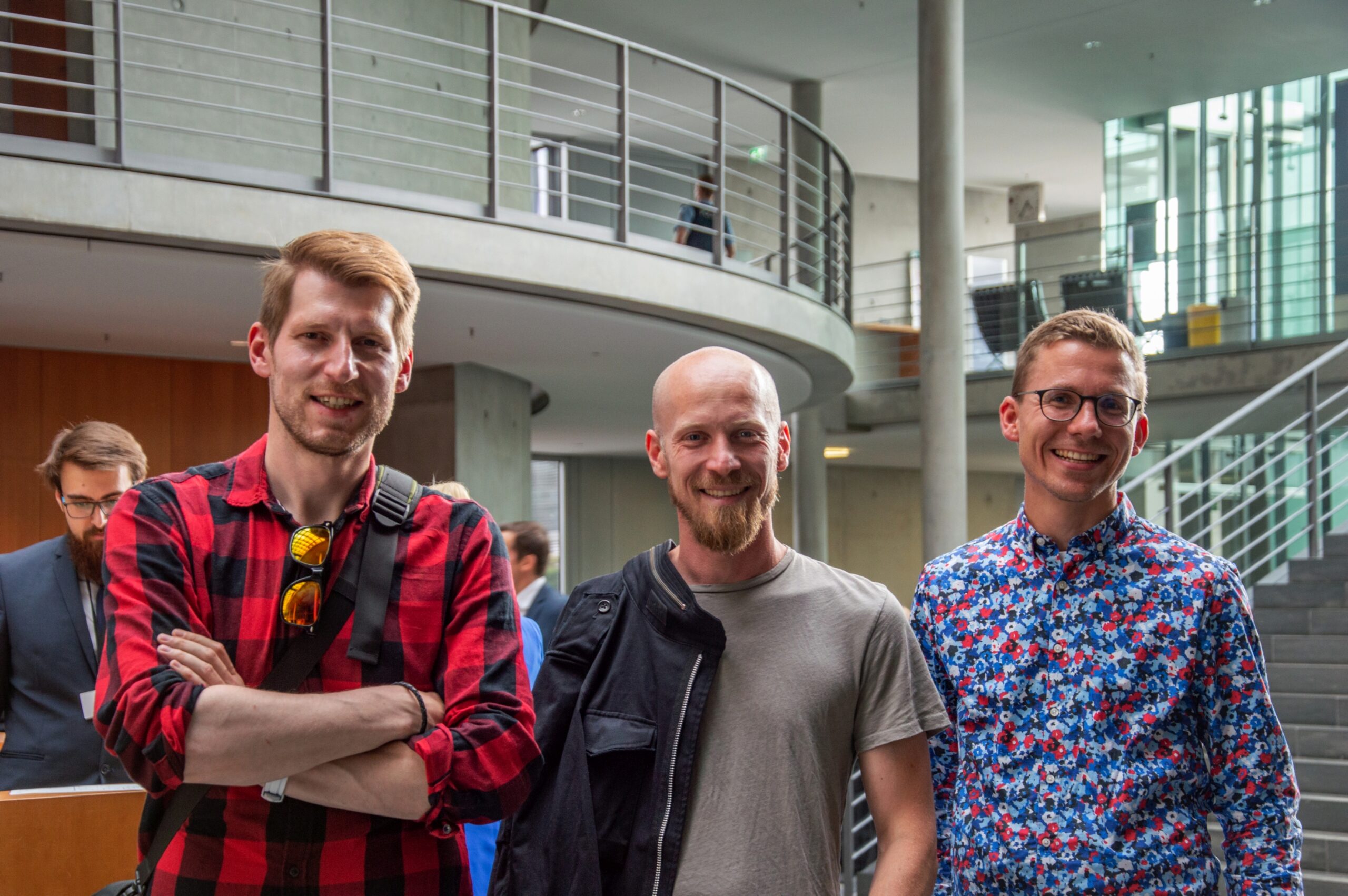 Drei lächelnde Männer stehen in einem Gebäude der Hochschule Coburg zusammen. Einer trägt ein rot kariertes Hemd, ein anderer ein graues T-Shirt und der dritte ein Hemd mit Blumenmuster. Im Hintergrund sind eine moderne Treppe und ein Geländer zu sehen, die zur lebhaften Atmosphäre ihres Treffens beitragen.