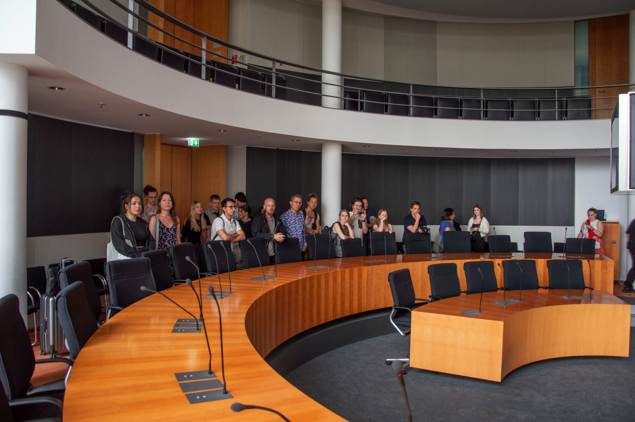 Eine Gruppe von Menschen steht in einem halbrunden Konferenzraum der Hochschule Coburg mit einem großen hölzernen Konferenztisch und schwarzen Stühlen. Der Raum hat hohe Decken und ein modernes Design mit glatter Wandverkleidung. Einige Personen blicken nach vorne.
