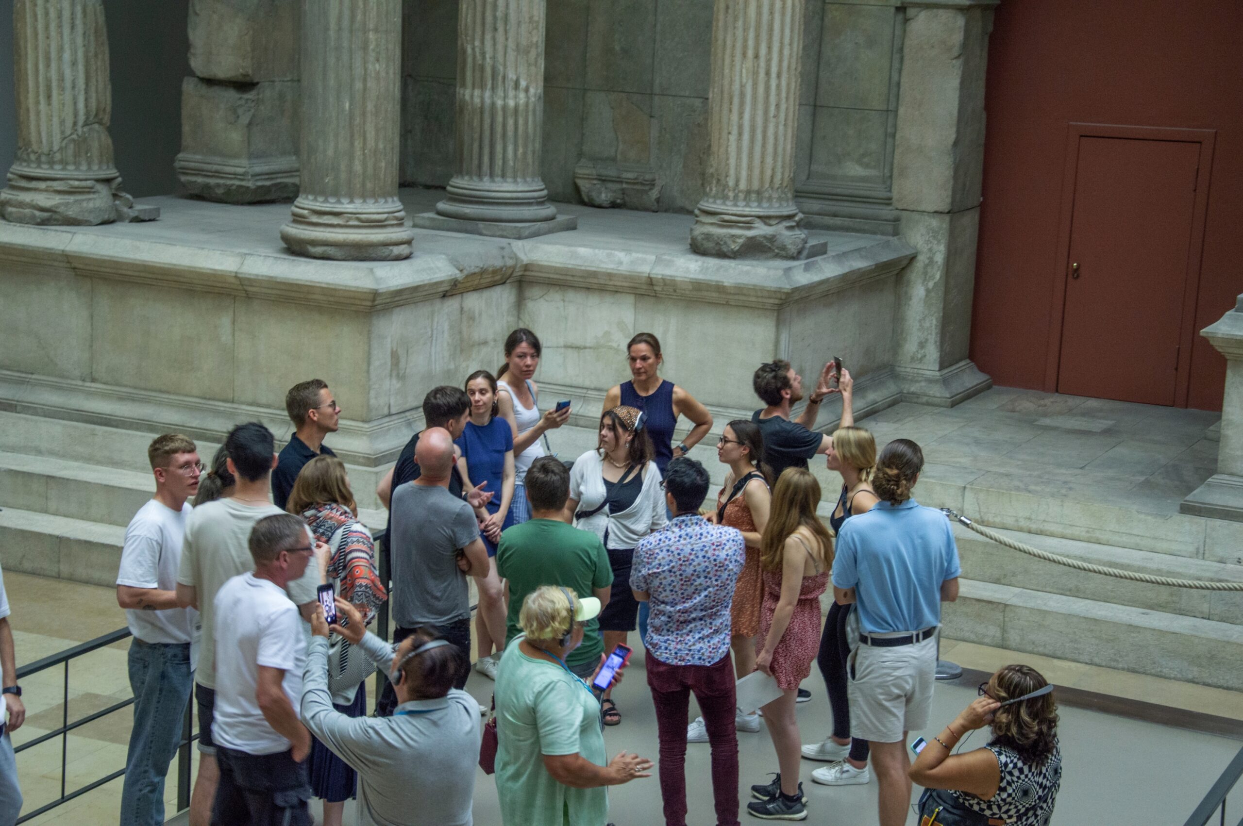 Eine Gruppe von Menschen versammelt sich in der Nähe alter Steinsäulen in einem Museum der Hochschule Coburg. Einige Personen machen Fotos, während andere sich unterhalten. Die Architektur zeichnet sich durch hohe Säulen und einen Steinmauerhintergrund aus und verbindet Geschichte mit der lebendigen Atmosphäre der Wissenschaft.
