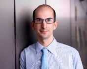 Ein Mann mit Brille, hellblauem Hemd und passender Krawatte steht vor einer reflektierenden Oberfläche an der Hochschule Coburg. Er blickt direkt in die Kamera und lächelt dabei leicht.