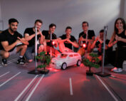 Eine Gruppe von sieben Personen der Hochschule Coburg posiert um ein kleines Modellauto auf einer Rennstrecke. Der Bereich ist mit roten Lichtern beleuchtet, in der Nähe stehen Miniaturbäume und Straßenlaternen. Die Gruppe lächelt und macht Handbewegungen und fängt einen Moment der Kameradschaft und Kreativität ein.