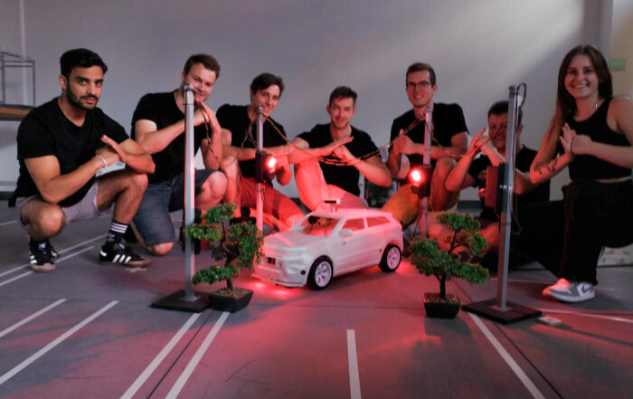 Eine Gruppe von sieben Personen der Hochschule Coburg posiert um ein kleines Modellauto auf einer Rennstrecke. Der Bereich ist mit roten Lichtern beleuchtet, in der Nähe stehen Miniaturbäume und Straßenlaternen. Die Gruppe lächelt und macht Handbewegungen und fängt einen Moment der Kameradschaft und Kreativität ein.