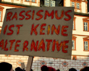 Eine protestierende Menschenmenge steht vor der historischen Hochschule Coburg und hält ein Pappschild mit der Aufschrift „Rassismus ist keine Alternative“. Der Himmel ist klar und hebt die kunstvollen architektonischen Details des Gebäudes hervor.
