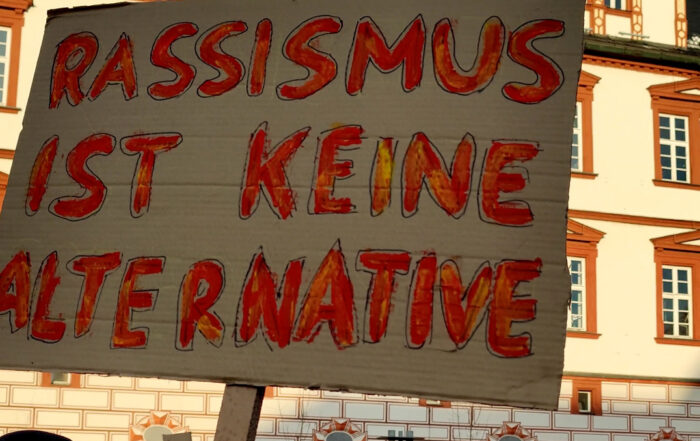 Eine protestierende Menschenmenge steht vor der historischen Hochschule Coburg und hält ein Pappschild mit der Aufschrift „Rassismus ist keine Alternative“. Der Himmel ist klar und hebt die kunstvollen architektonischen Details des Gebäudes hervor.