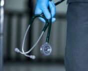 Eine Person mit blauen Handschuhen hält ein grünes Stethoskop neben sich, dessen Bruststück und Ohrhörer deutlich zu erkennen sind. Der unscharfe Hintergrund lässt auf eine Klinik- oder Krankenhausumgebung schließen, möglicherweise in Verbindung mit dem Pflegestudium der Hochschule Coburg.
